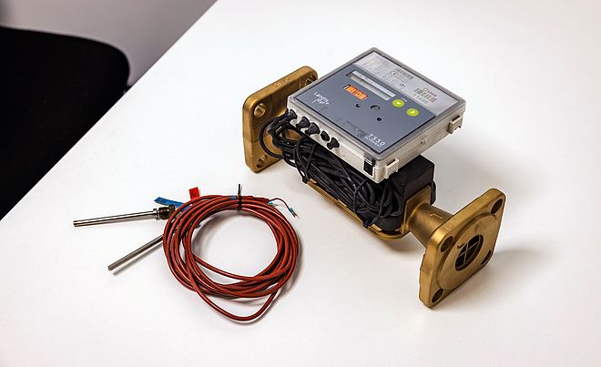 Auf einem weißen Tisch liegt ein Durchflussmessgerät mit zwei massiven, goldfarbenen Flanschen. Auf dem Gerät ist ein elektronisches Modul mit Display und Bedientasten montiert, darunter sind schwarze Kabel ordentlich aufgewickelt. Daneben befindet sich ein Bündel roter Sensorkabel mit zwei Metallfühlern. Die Szene zeigt Messtechnik für Energie- oder Wärmenetze. Verlinkung zur Messstellenbetrieb Wärme Seite.