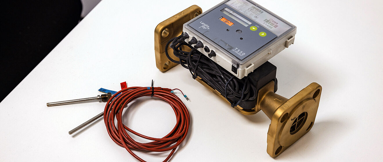 Auf einem weißen Tisch liegt ein Durchflussmessgerät mit zwei massiven, goldfarbenen Flanschen. Auf dem Gerät ist ein elektronisches Modul mit Display und Bedientasten montiert, darunter sind schwarze Kabel ordentlich aufgewickelt. Daneben befindet sich ein Bündel roter Sensorkabel mit zwei Metallfühlern. Die Szene zeigt Messtechnik für Energie- oder Wärmenetze. Verlinkung zur Messstellenbetrieb Wärme Seite.