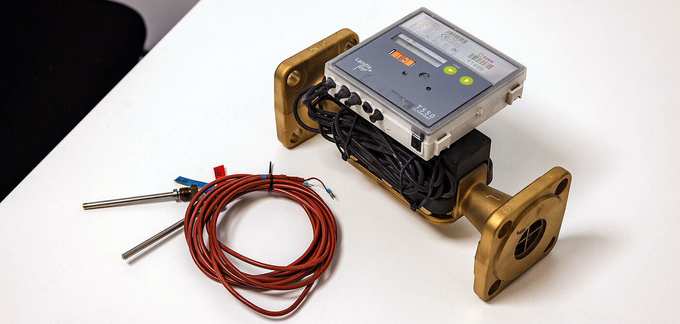 Auf einem weißen Tisch liegt ein Durchflussmessgerät mit zwei massiven, goldfarbenen Flanschen. Auf dem Gerät ist ein elektronisches Modul mit Display und Bedientasten montiert, darunter sind schwarze Kabel ordentlich aufgewickelt. Daneben befindet sich ein Bündel roter Sensorkabel mit zwei Metallfühlern. Die Szene zeigt Messtechnik für Energie- oder Wärmenetze. Verlinkung zur Messstellenbetrieb Wärme Seite.