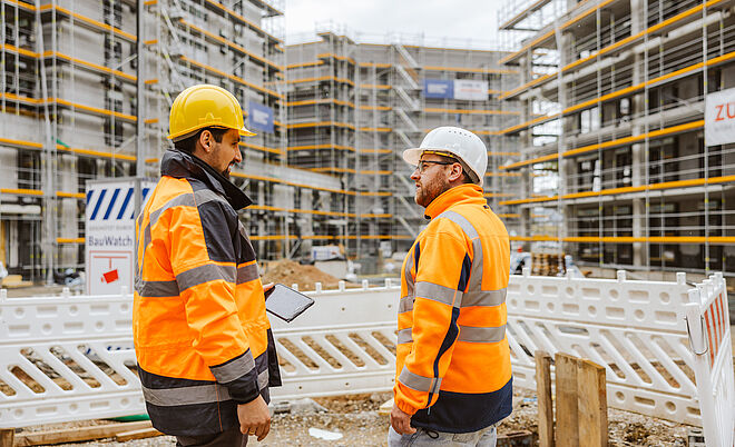Zwei Personen in orangefarbener Warnschutzkleidung und Schutzhelmen stehen auf einer Baustelle und sprechen miteinander. Eine Person hält ein Tablet in der Hand. Im Hintergrund sind mehrere Gebäude im Rohbau mit Gerüsten und gelben Sicherheitsleisten zu sehen. Vor den Personen befindet sich eine weiße Baustellenabsperrung, dahinter liegen Erdmaterialien. Verlinkung zur Hausanschluss_Kunde Seite.