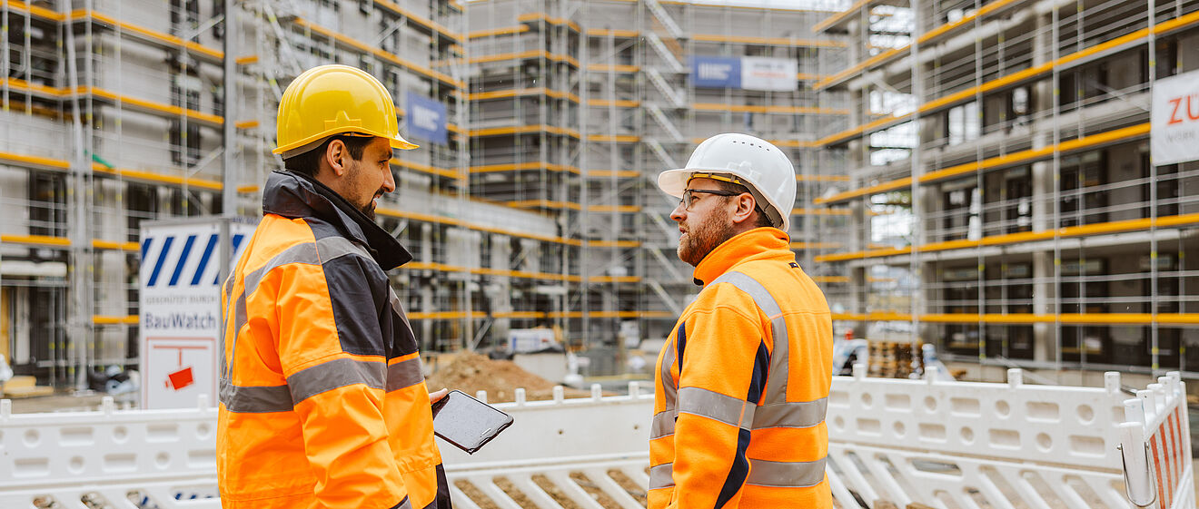 Zwei Personen in orangefarbener Warnschutzkleidung und Schutzhelmen stehen auf einer Baustelle und sprechen miteinander. Eine Person hält ein Tablet in der Hand. Im Hintergrund sind mehrere Gebäude im Rohbau mit Gerüsten und gelben Sicherheitsleisten zu sehen. Vor den Personen befindet sich eine weiße Baustellenabsperrung, dahinter liegen Erdmaterialien. Verlinkung zur Hausanschluss_Kunde Seite.