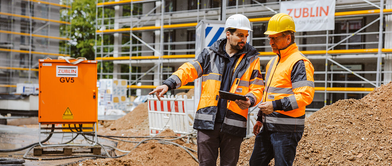 Zwei Personen in Sicherheitskleidung und Helmen stehen auf einer Baustelle vor einem großen Erdhaufen. Beide tragen orange Warnjacken mit reflektierenden Streifen. Eine Person hält ein Klemmbrett, die andere zeigt mit der Hand in eine bestimmte Richtung, offenbar zur Besprechung der Arbeiten. Im Hintergrund sind Gerüste an einem Gebäude sowie ein orangefarbener Baustromverteiler mit der Aufschrift ‚ZÜBLIN GVS‘ zu sehen. Die Szene verdeutlicht Bauleitung und Koordination auf einer Baustelle. Verlinkung zur Störung melden Seite.