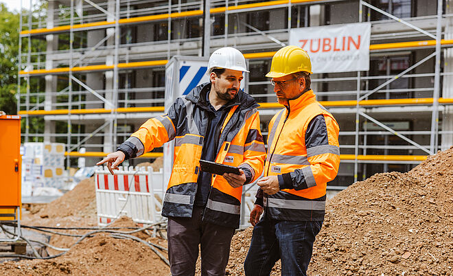 Zwei Personen in Sicherheitskleidung und Helmen stehen auf einer Baustelle vor einem großen Erdhaufen. Beide tragen orange Warnjacken mit reflektierenden Streifen. Eine Person hält ein Klemmbrett, die andere zeigt mit der Hand in eine bestimmte Richtung, offenbar zur Besprechung der Arbeiten. Im Hintergrund sind Gerüste an einem Gebäude sowie ein orangefarbener Baustromverteiler mit der Aufschrift ‚ZÜBLIN GVS‘ zu sehen. Die Szene verdeutlicht Bauleitung und Koordination auf einer Baustelle. Verlinkung zur Störung melden Seite.