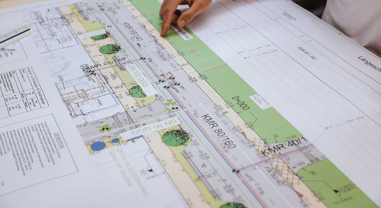 Detailaufnahme eines technischen Plans für Leitungsverlegung. Auf dem Tisch liegt eine großformatige Bauzeichnung mit farbigen Markierungen, Linien und Beschriftungen. Der Plan zeigt eine Straßenansicht mit Grünflächen, Leitungsführungen und technischen Daten wie Rohrdimensionen (z. B. KMR 80/160, KMR 40/1). Eine Hand deutet auf einen bestimmten Bereich des Plans, um eine Position oder Messung hervorzuheben. Im Hintergrund sind weitere zusammengerollte Pläne sichtbar. Die Szene verdeutlicht präzise Planung und Dokumentation für Infrastrukturprojekte.
