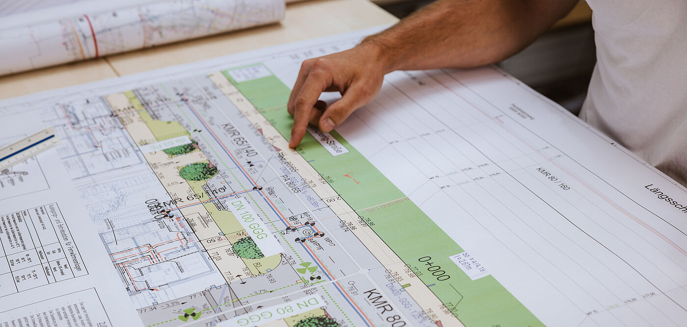 Detailaufnahme eines technischen Plans für Leitungsverlegung. Auf dem Tisch liegt eine großformatige Bauzeichnung mit farbigen Markierungen, Linien und Beschriftungen. Der Plan zeigt eine Straßenansicht mit Grünflächen, Leitungsführungen und technischen Daten wie Rohrdimensionen (z. B. KMR 80/160, KMR 40/1). Eine Hand deutet auf einen bestimmten Bereich des Plans, um eine Position oder Messung hervorzuheben. Im Hintergrund sind weitere zusammengerollte Pläne sichtbar. Die Szene verdeutlicht präzise Planung und Dokumentation für Infrastrukturprojekte.