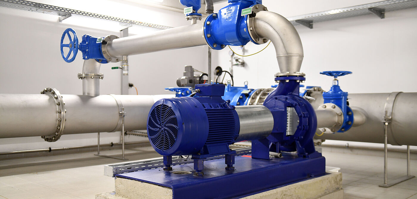 Industriepumpe in einer technischen Anlage: Im Vordergrund steht eine große, blau lackierte Kreiselpumpe mit Elektromotor, montiert auf einem Betonfundament. Die Pumpe ist über dicke, silberfarbene Rohrleitungen mit weiteren Armaturen verbunden. Mehrere blaue Absperrventile und Rohrbögen sind sichtbar. Die Umgebung ist ein heller, sauberer Technikraum mit gefliestem Boden und Kabeltrassen an der Wand.