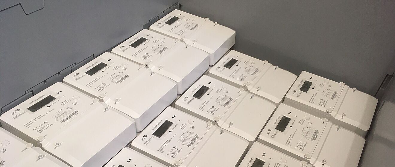 In einer großen Kunststoffbox liegen mehrere weiße digitale Stromzähler ordentlich gestapelt. Jeder Zähler verfügt über ein rechteckiges Display, Bedientasten und technische Beschriftungen. Die Geräte sind für den Einsatz in modernen Stromnetzen vorgesehen und zeigen eine einheitliche, kompakte Bauform. Verlinkung zur digitale Stromzähler Seite.