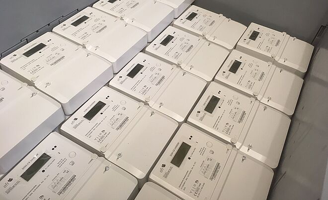 In einer großen Kunststoffbox liegen mehrere weiße digitale Stromzähler ordentlich gestapelt. Jeder Zähler verfügt über ein rechteckiges Display, Bedientasten und technische Beschriftungen. Die Geräte sind für den Einsatz in modernen Stromnetzen vorgesehen und zeigen eine einheitliche, kompakte Bauform. Verlinkung zur digitale Stromzähler Seite.