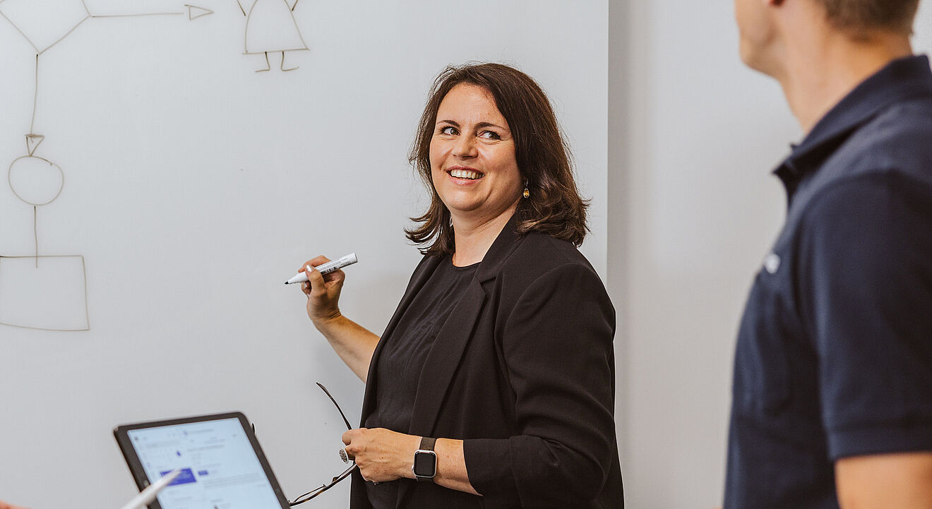 Drei Personen stehen an einem runden Tisch in einem Besprechungsraum. Auf dem Tisch liegen ein Tablet und ein geschlossenes Notebook. Im Hintergrund befindet sich ein großes Whiteboard, auf dem ein einfaches Diagramm mit Pfeilen und geometrischen Formen sowie eine kleine Figur gezeichnet sind. Eine Person schreibt mit einem Marker auf das Whiteboard, während die anderen beiden aufmerksam zuschauen. Die Umgebung wirkt modern und hell, mit weißer Wand und klarer Struktur.