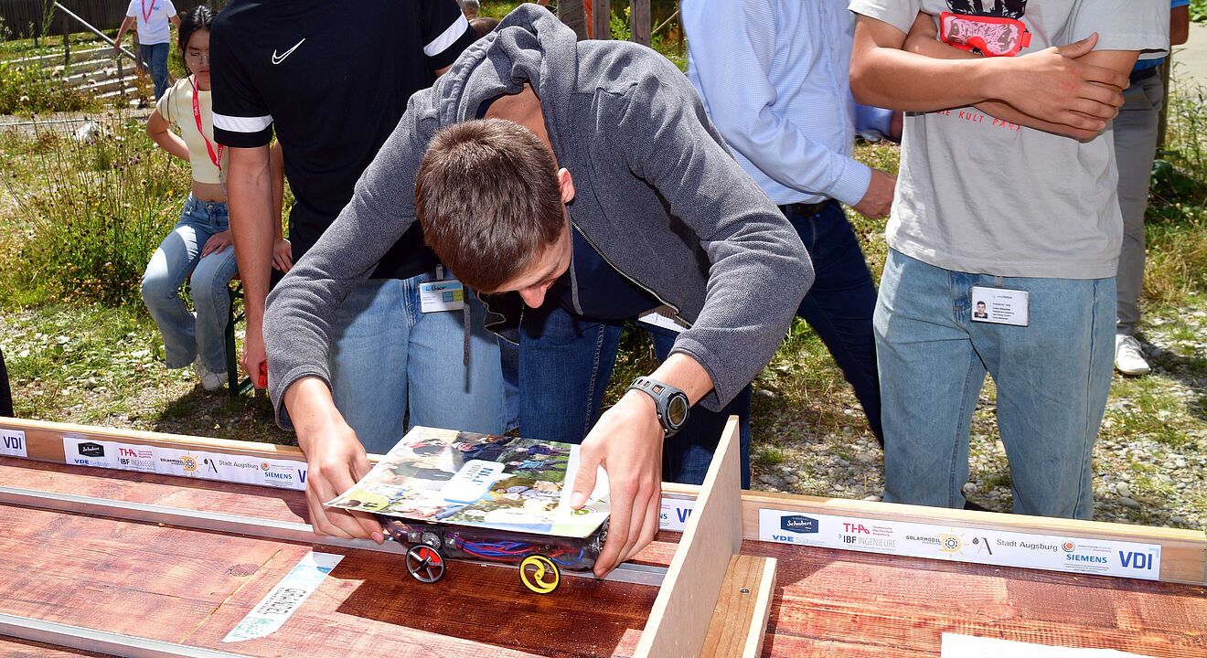 Mehrere Personen stehen im Freien an einem Tisch und bereiten ein kleines Fahrzeug für einen Wettbewerb vor. Das Fahrzeug ist auf einer Holzbahn platziert, die mit Logos verschiedener Partner wie VDI und TUM versehen ist. Auf dem Tisch liegen Bauteile und Werkzeuge. Im Hintergrund sind Bäume, ein Zaun und weitere Personen sichtbar.