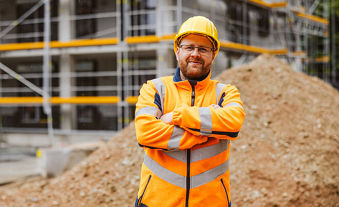 Eine Person trägt eine leuchtend orangefarbene Warnschutzjacke mit reflektierenden Streifen und einen gelben Schutzhelm. Die Person steht mit verschränkten Armen vor einer Baustelle. Im Hintergrund sind ein großer Erdhaufen und ein Gerüst mit gelben Sicherheitsleisten zu sehen. Verlinkung zur swa Netze investieren ins Versorgungsnetz Seite.