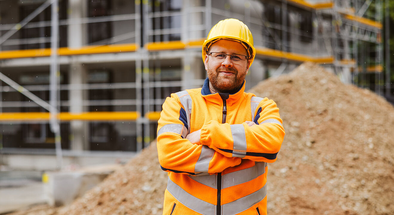 Eine Person trägt eine leuchtend orangefarbene Warnschutzjacke mit reflektierenden Streifen und einen gelben Schutzhelm. Die Person steht mit verschränkten Armen vor einer Baustelle. Im Hintergrund sind ein großer Erdhaufen und ein Gerüst mit gelben Sicherheitsleisten zu sehen. Verlinkung zur swa Netze investieren ins Versorgungsnetz Seite.
