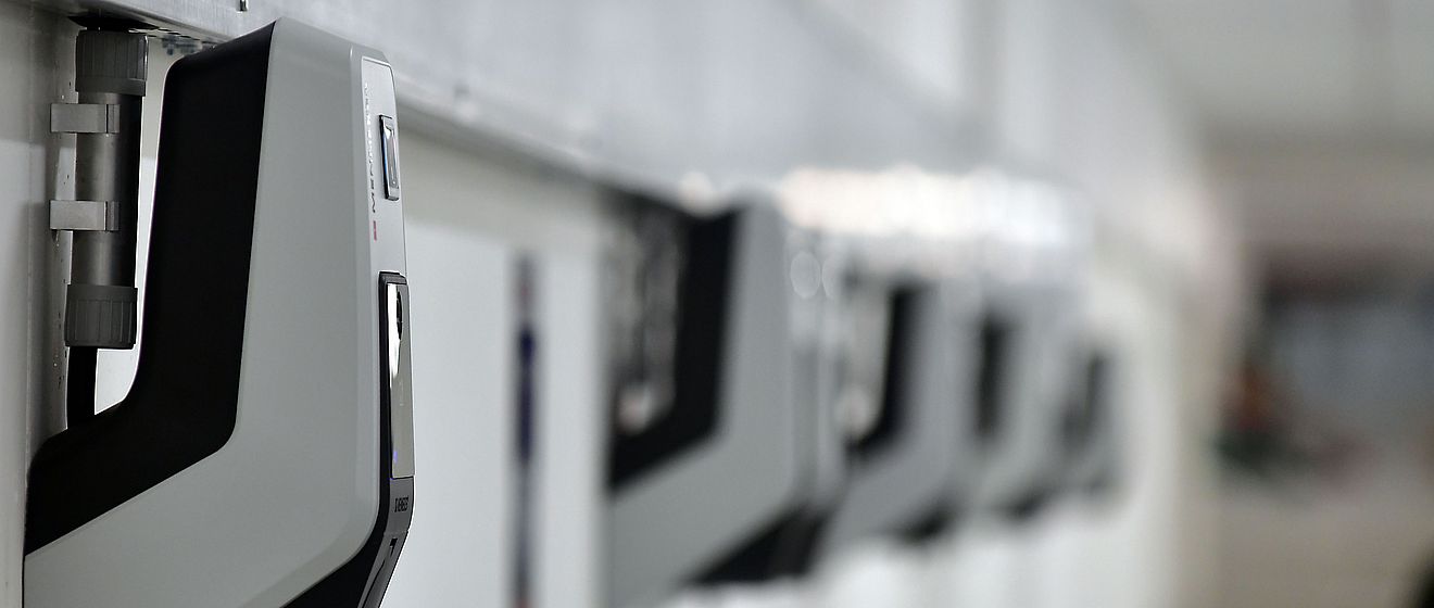 Reihe von Wandladestationen für Elektrofahrzeuge in einem Parkhaus oder einer Garage: Mehrere moderne, kompakte Ladegeräte sind an einer weißen Wand montiert und gleichmäßig angeordnet. Die Geräte sind grau-schwarz und über Kabelkanäle verbunden. Die Szene symbolisiert Ladeinfrastruktur für Elektromobilität. Verlinkung zur eMobilität Seite.