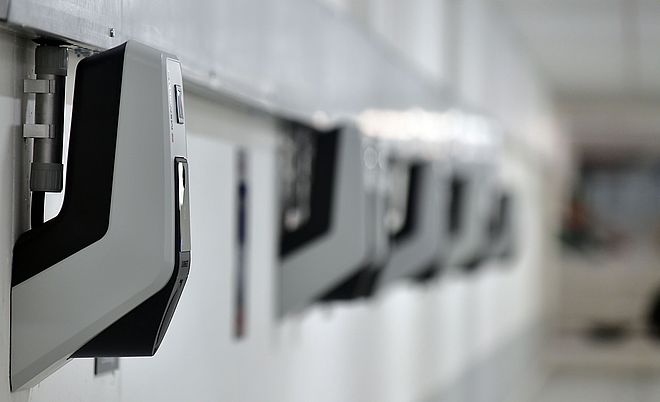 Reihe von Wandladestationen für Elektrofahrzeuge in einem Parkhaus oder einer Garage: Mehrere moderne, kompakte Ladegeräte sind an einer weißen Wand montiert und gleichmäßig angeordnet. Die Geräte sind grau-schwarz und über Kabelkanäle verbunden. Die Szene symbolisiert Ladeinfrastruktur für Elektromobilität. Verlinkung zur eMobilität Seite.