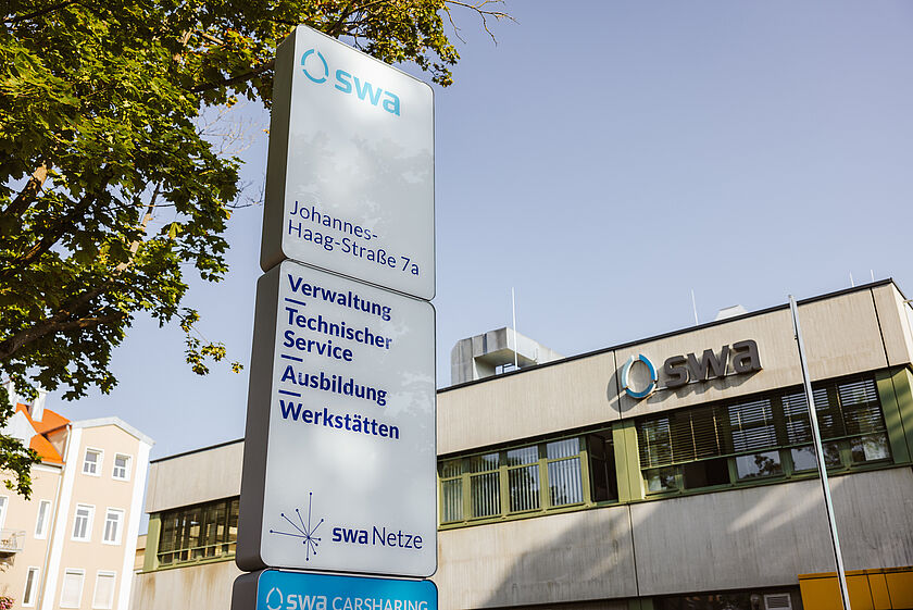 Gebäude der swa Netze GmbH an der Johannes-Haag-Straße 7a in Augsburg. Im Vordergrund steht ein großes Hinweisschild mit dem swa-Logo und den Angaben: ‚Johannes-Haag-Straße 7a‘ sowie den Bereichen Verwaltung, Technischer Service, Ausbildung und Werkstätten. Darunter befindet sich das Logo von swa Netze und ein Hinweis auf swa Carsharing. Im Hintergrund ist das Verwaltungsgebäude mit dem swa-Logo an der Fassade zu sehen. Links ragen einige Wohnhäuser und ein Baum ins Bild, der für Schatten sorgt. Der Himmel ist klar und sonnig.
