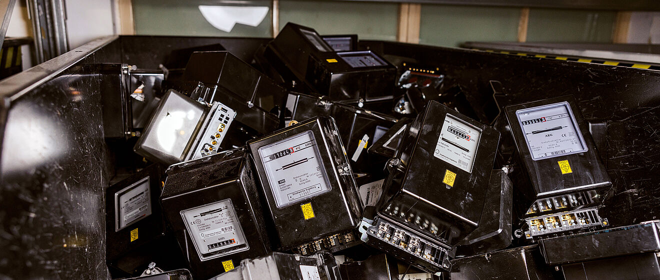 Ein Behälter ist mit zahlreichen ausgebauten, schwarzen Stromzählern gefüllt. Die Geräte sind rechteckig, haben ein Display mit Skalen und Zahlen sowie Anschlussklemmen an der Unterseite. Einige Zähler sind sichtbar beschädigt oder zerkratzt. Im Hintergrund sind weitere Zähler und ein Regal zu erkennen. Die Szene zeigt ausrangierte Messtechnik, vermutlich zur Entsorgung oder zum Recycling. Verlinkung zur Zählerentfernung Seite.