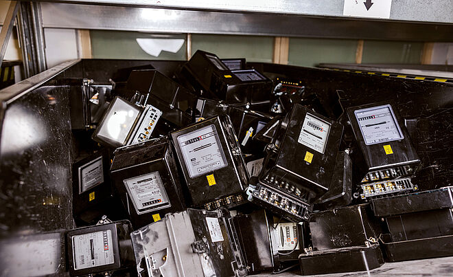 Ein Behälter ist mit zahlreichen ausgebauten, schwarzen Stromzählern gefüllt. Die Geräte sind rechteckig, haben ein Display mit Skalen und Zahlen sowie Anschlussklemmen an der Unterseite. Einige Zähler sind sichtbar beschädigt oder zerkratzt. Im Hintergrund sind weitere Zähler und ein Regal zu erkennen. Die Szene zeigt ausrangierte Messtechnik, vermutlich zur Entsorgung oder zum Recycling. Verlinkung zur Zählerentfernung Seite.