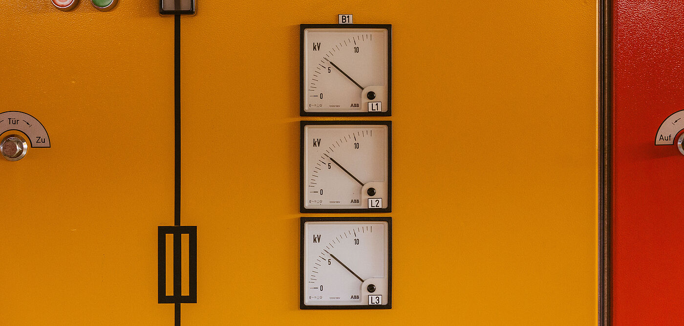 Detailansicht einer gelben Schaltanlage mit drei analogen Messinstrumenten für Hochspannung: Die Anzeigen zeigen Werte in Kilovolt (kV) für die Phasen L1, L2 und L3. Links sind zwei Drucktaster in Grün und Rot sowie ein Schalter sichtbar. Schwarze Linien und Symbole auf der Front markieren die elektrische Schaltung. Die Szene zeigt eine Steuer- und Überwachungseinheit für Energieverteilung in einem Technikraum.