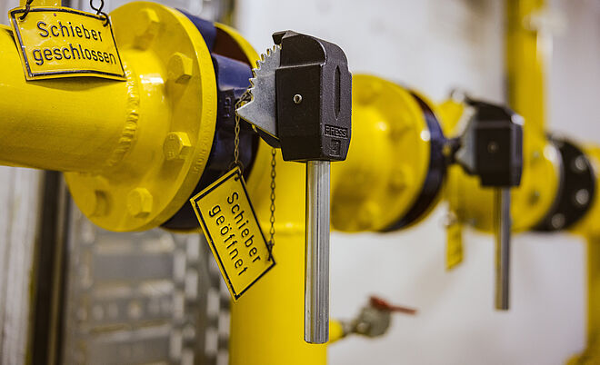 Detailaufnahme einer gelben Rohrleitungsarmatur mit zwei Schiebern, die jeweils mit Metallschlüsseln gesichert sind. An den Schiebern hängen gelbe Schilder mit der Aufschrift ‚Schieber geschlossen‘ und ‚Schieber geöffnet‘. Im Hintergrund sind weitere gelbe Rohrverbindungen und metallische Komponenten sichtbar. Verlinkung zur Netzzugang Gas Seite.