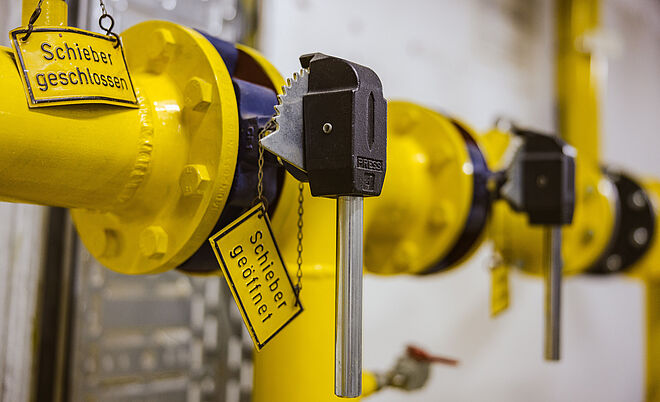 Detailaufnahme einer gelben Rohrleitungsarmatur mit zwei Schiebern, die jeweils mit Metallschlüsseln gesichert sind. An den Schiebern hängen gelbe Schilder mit der Aufschrift ‚Schieber geschlossen‘ und ‚Schieber geöffnet‘. Im Hintergrund sind weitere gelbe Rohrverbindungen und metallische Komponenten sichtbar. Verlinkung zur Netzzugang Gas Seite.