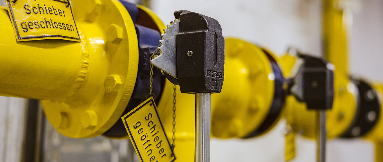 Detailaufnahme einer gelben Rohrleitungsarmatur mit zwei Schiebern, die jeweils mit Metallschlüsseln gesichert sind. An den Schiebern hängen gelbe Schilder mit der Aufschrift ‚Schieber geschlossen‘ und ‚Schieber geöffnet‘. Im Hintergrund sind weitere gelbe Rohrverbindungen und metallische Komponenten sichtbar. Verlinkung zur Netzzugang Gas Seite.
