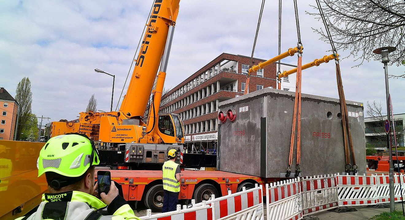 Straßenszene mit Baustelle im urbanen Umfeld: Ein großer gelber Mobilkran hebt einen massiven, rechteckigen Beton-Schacht (Herstelleraufschrift „PRIES“) mit mehreren angeschlagenen Hebegurten von einem Tieflader. Der Arbeitsbereich ist durch rot‑weiße Absperrgitter gesichert. Mehrere Personen in hochsichtbarer Schutzkleidung und Helmen beaufsichtigen den Hebevorgang; eine Person im Vordergrund fotografiert mit einem Smartphone. Im Hintergrund steht ein mehrgeschossiges Backsteingebäude mit Balkonen; an der Fassade ist „GUDRUNS APOTHEKE“ erkennbar. Bewölkter Himmel, Baum und St„Kraßenbeleuchtung rahmen die Szene. Verlinkung zur Seite Koppelstation Wolframviertel.