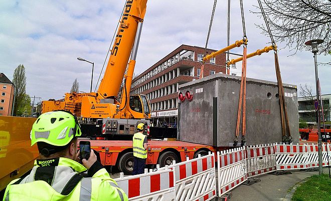 Straßenszene mit Baustelle im urbanen Umfeld: Ein großer gelber Mobilkran hebt einen massiven, rechteckigen Beton-Schacht (Herstelleraufschrift „PRIES“) mit mehreren angeschlagenen Hebegurten von einem Tieflader. Der Arbeitsbereich ist durch rot‑weiße Absperrgitter gesichert. Mehrere Personen in hochsichtbarer Schutzkleidung und Helmen beaufsichtigen den Hebevorgang; eine Person im Vordergrund fotografiert mit einem Smartphone. Im Hintergrund steht ein mehrgeschossiges Backsteingebäude mit Balkonen; an der Fassade ist „GUDRUNS APOTHEKE“ erkennbar. Bewölkter Himmel, Baum und St„Kraßenbeleuchtung rahmen die Szene. Verlinkung zur Seite Koppelstation Wolframviertel.