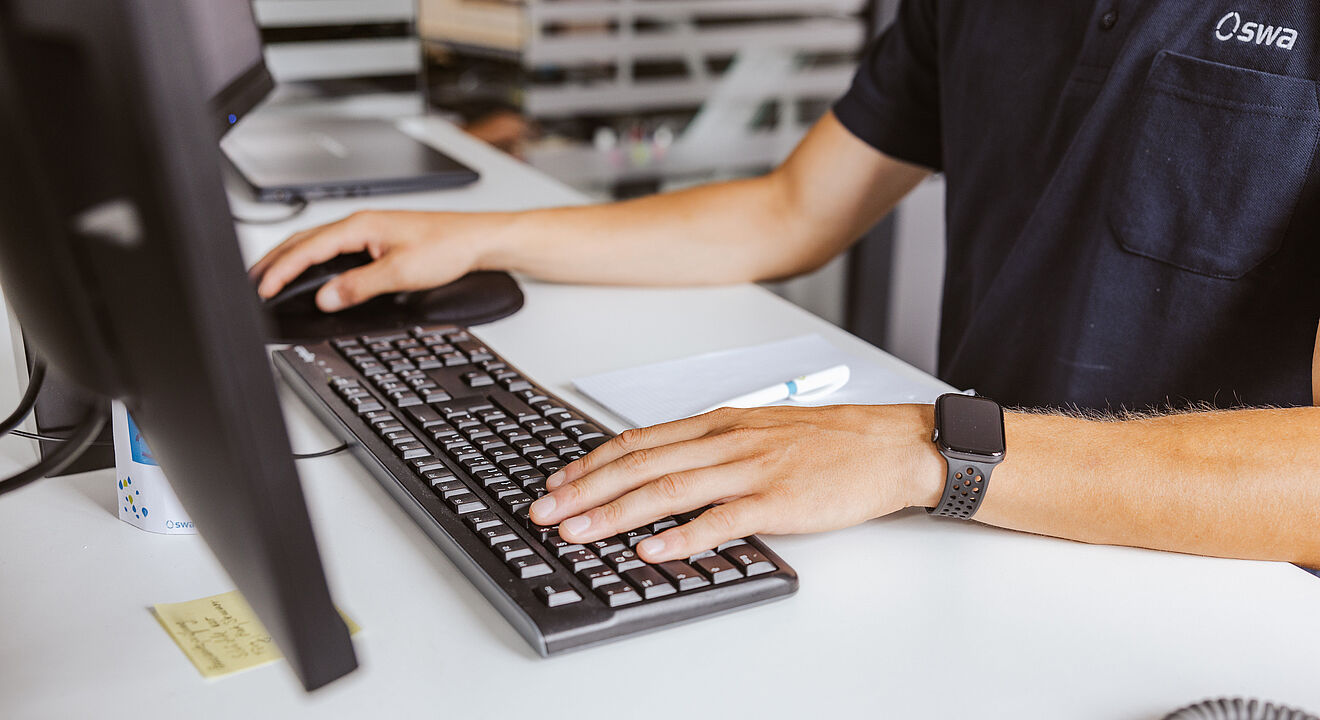 Eine Person sitzt an einem Schreibtisch und arbeitet an einem Computer. Die Hände liegen auf einer Tastatur, daneben befindet sich eine Computermaus. Auf dem Schreibtisch liegen ein Notizblock, ein Stift, ein Telefonhörer und ein Post-it-Zettel. Die Person trägt ein dunkles Poloshirt mit dem Logo „swa“ und eine Smartwatch am Handgelenk. Im Hintergrund sind unscharf Regale mit Unterlagen zu erkennen.