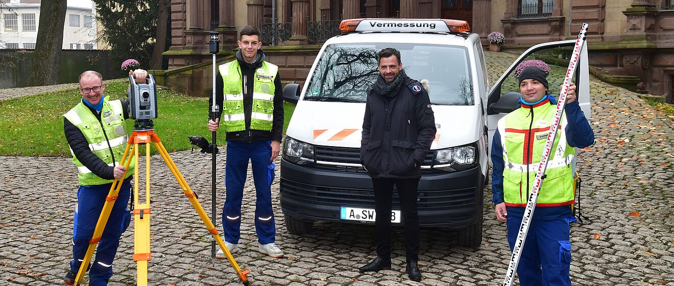 Vier Personen in Warnwesten stehen vor einem weißen Vermessungsfahrzeug mit der Aufschrift ‚Vermessung‘. Sie befinden sich auf einem gepflasterten Platz vor einem historischen Gebäude mit Säulen und großen Fenstern. Links ist ein gelbes Stativ mit einem Vermessungsgerät zu sehen, daneben hält eine Person eine Messlatte, und eine weitere Person trägt ein GPS-Messgerät. Rechts steht ein Transporter mit orangefarbenen Warnmarkierungen. Die Szene zeigt ein Team bei der Vermessungsarbeit im Außenbereich. Verlinkung zur Vemessung Seite.