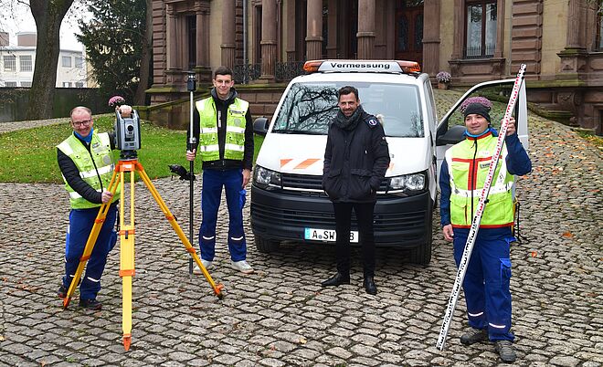 Vier Personen in Warnwesten stehen vor einem weißen Vermessungsfahrzeug mit der Aufschrift ‚Vermessung‘. Sie befinden sich auf einem gepflasterten Platz vor einem historischen Gebäude mit Säulen und großen Fenstern. Links ist ein gelbes Stativ mit einem Vermessungsgerät zu sehen, daneben hält eine Person eine Messlatte, und eine weitere Person trägt ein GPS-Messgerät. Rechts steht ein Transporter mit orangefarbenen Warnmarkierungen. Die Szene zeigt ein Team bei der Vermessungsarbeit im Außenbereich. Verlinkung zur Vemessung Seite.