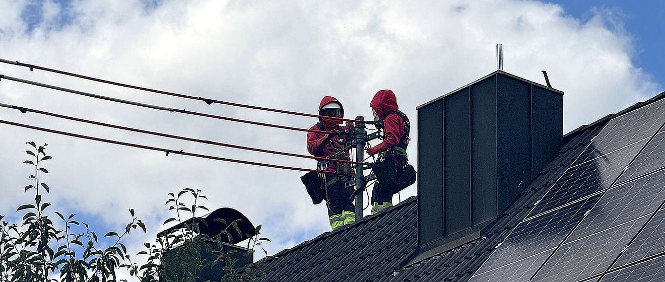 Zwei mit persönlicher Schutzausrüstung und Absturzsicherung ausgestattete Einsatzkräfte arbeiten auf dem Dach eines Wohngebäudes an einer Freileitung. Auf der rechten Dachseite sind großflächige Photovoltaikmodule installiert. Im Vordergrund sind Baumkronen sichtbar, im Hintergrund ein bewölkter Himmel. Das Bild verdeutlicht Arbeiten an der Infrastruktur im Umfeld moderner Energieerzeugung. Verlinkung zur Freileitungsisolierung Seite.