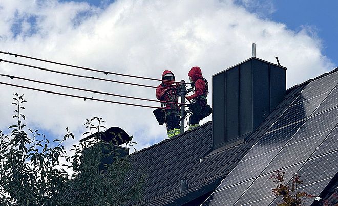 Zwei mit persönlicher Schutzausrüstung und Absturzsicherung ausgestattete Einsatzkräfte arbeiten auf dem Dach eines Wohngebäudes an einer Freileitung. Auf der rechten Dachseite sind großflächige Photovoltaikmodule installiert. Im Vordergrund sind Baumkronen sichtbar, im Hintergrund ein bewölkter Himmel. Das Bild verdeutlicht Arbeiten an der Infrastruktur im Umfeld moderner Energieerzeugung. Verlinkung zur Freileitungsisolierung Seite.