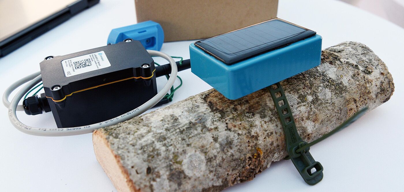 Auf einem weißen Tisch liegt ein Stück Holz, an dem ein blaues Sensormodul mit Solarpanel befestigt ist. Daneben befindet sich ein schwarzes elektronisches Gerät mit Kabelanschluss und QR-Code sowie ein weiteres kleines blaues Bauteil. Im Hintergrund steht ein brauner Karton. Die Geräte sind Teil einer IoT-Lösung zur Umwelt- oder Zustandsüberwachung.