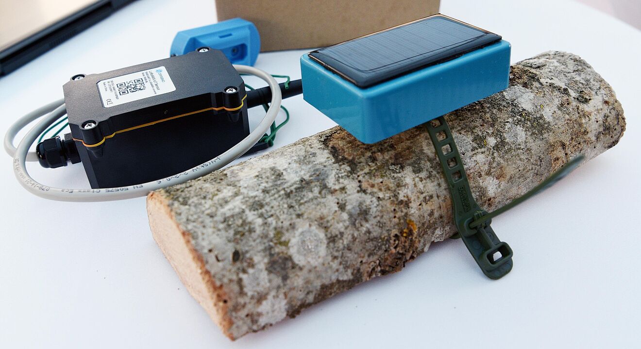 Auf einem weißen Tisch liegt ein Stück Holz, an dem ein blaues Sensormodul mit Solarpanel befestigt ist. Daneben befindet sich ein schwarzes elektronisches Gerät mit Kabelanschluss und QR-Code sowie ein weiteres kleines blaues Bauteil. Im Hintergrund steht ein brauner Karton. Die Geräte sind Teil einer IoT-Lösung zur Umwelt- oder Zustandsüberwachung.
