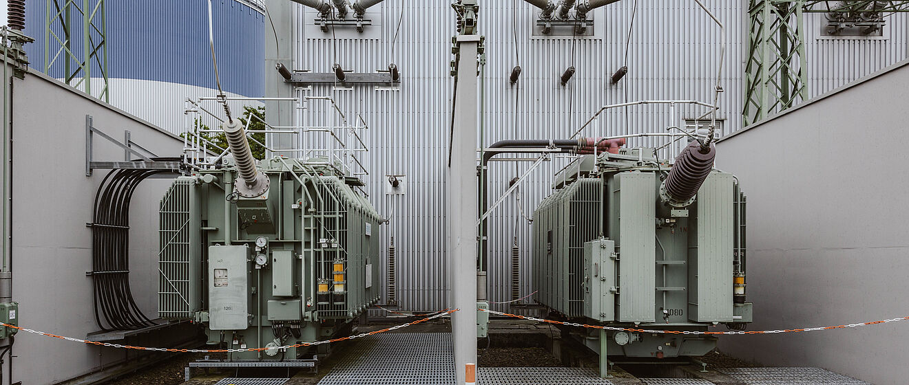 Zwei große Hochspannungstransformatoren in einem Umspannwerk: Die Transformatoren sind grau lackiert und stehen nebeneinander auf Gitterrosten, umgeben von Schutzgeländern und Rohrleitungen. Über den Transformatoren verlaufen mehrere Isolatoren und Anschlussleitungen, die an die Gebäudewand angeschlossen sind. Im Hintergrund sind eine Metallfassade, Kabeldurchführungen und ein Stahlgittermast sichtbar. Die Szene zeigt zentrale Komponenten der Energieübertragung und Netztechnik. Verlinkung zur Auswahl Netzzugänge Seite.
