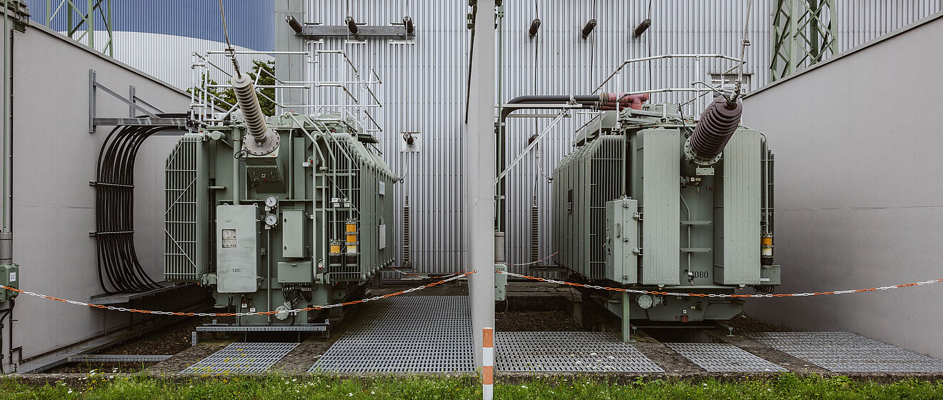 Zwei große Hochspannungstransformatoren in einem Umspannwerk: Die Transformatoren sind grau lackiert und stehen nebeneinander auf Gitterrosten, umgeben von Schutzgeländern und Rohrleitungen. Über den Transformatoren verlaufen mehrere Isolatoren und Anschlussleitungen, die an die Gebäudewand angeschlossen sind. Im Hintergrund sind eine Metallfassade, Kabeldurchführungen und ein Stahlgittermast sichtbar. Die Szene zeigt zentrale Komponenten der Energieübertragung und Netztechnik. Verlinkung zur Auswahl Netzzugänge Seite.