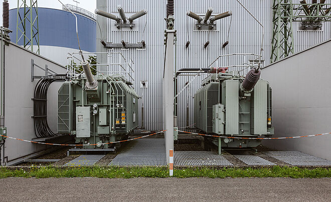 Zwei große Hochspannungstransformatoren in einem Umspannwerk: Die Transformatoren sind grau lackiert und stehen nebeneinander auf Gitterrosten, umgeben von Schutzgeländern und Rohrleitungen. Über den Transformatoren verlaufen mehrere Isolatoren und Anschlussleitungen, die an die Gebäudewand angeschlossen sind. Im Hintergrund sind eine Metallfassade, Kabeldurchführungen und ein Stahlgittermast sichtbar. Die Szene zeigt zentrale Komponenten der Energieübertragung und Netztechnik. Verlinkung zur Auswahl Netzzugänge Seite.
