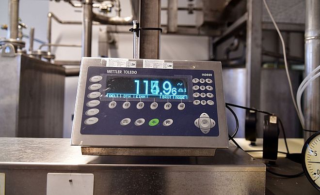 Nahaufnahme einer Edelstahlwaage in einer industriellen Prozessumgebung. Auf einem digitalen Anzeigegerät der Marke „Mettler Toledo“ ist ein Messwert von rund 718,9 zu sehen. Die Waage steht vor einem Netzwerk aus Edelstahlrohren und technischen Anlagenteilen. Die Umgebung wirkt sauber, technisch und auf den Betrieb einer Versorgungs- oder Produktionsanlage ausgerichtet.