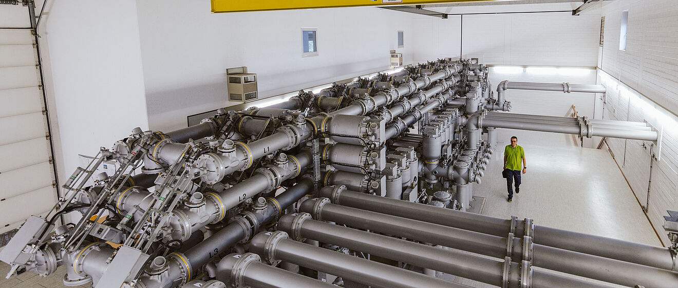 Innenansicht einer Schaltanlage in einem Umspannwerk: Mehrere große, silberfarbene Hochspannungs-Schaltrohre verlaufen parallel und sind mit Ventilen, Schaltern und Isolatoren ausgestattet. Die Anlage befindet sich in einer Halle mit Betonwänden und Decke, darüber ein gelber Kran für Wartungsarbeiten. Eine Person in Warnschutzkleidung geht entlang der Anlage. Die Szene zeigt moderne Gasisolierte Schalttechnik (GIS) für Stromversorgung und Netzbetrieb. Verlinkung zur Netzzugang Strom Seite.