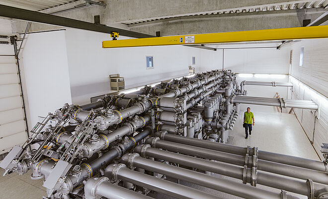 Innenansicht einer Schaltanlage in einem Umspannwerk: Mehrere große, silberfarbene Hochspannungs-Schaltrohre verlaufen parallel und sind mit Ventilen, Schaltern und Isolatoren ausgestattet. Die Anlage befindet sich in einer Halle mit Betonwänden und Decke, darüber ein gelber Kran für Wartungsarbeiten. Eine Person in Warnschutzkleidung geht entlang der Anlage. Die Szene zeigt moderne Gasisolierte Schalttechnik (GIS) für Stromversorgung und Netzbetrieb. Verlinkung zur Netzzugang Strom Seite.
