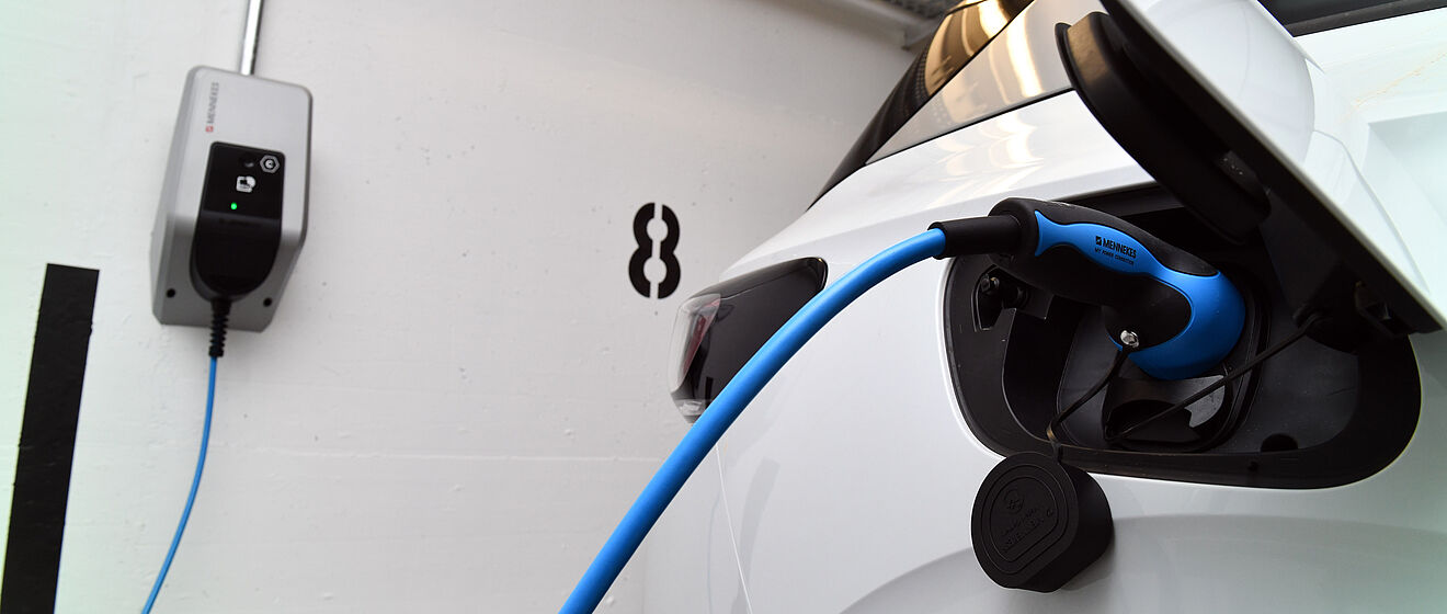 Elektroauto wird an einer Wandladestation geladen: Ein weißes Fahrzeug steht in einer Garage oder einem Parkhaus, das blaue Ladekabel ist in die geöffnete Ladebuchse des Autos eingesteckt. An der Wand befindet sich eine kompakte Ladestation mit LED-Anzeige. Die Szene symbolisiert nachhaltige Elektromobilität und Ladeinfrastruktur. Verlinkung zur eMobilität Seite.