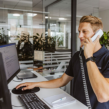 Eine Person sitzt an einem Schreibtisch und telefoniert mit einem Festnetztelefon. Die Person hält den Hörer am Ohr und bedient gleichzeitig die Computermaus. Vor der Person steht ein Monitor, auf dem eine Webseite oder ein Dokument geöffnet ist. Auf dem Schreibtisch liegen eine Tastatur, ein Notizblock mit Stift und ein Telefonhörer. Im Hintergrund sind Glaswände, Büroeinrichtung und grüne Pflanzen zu sehen.