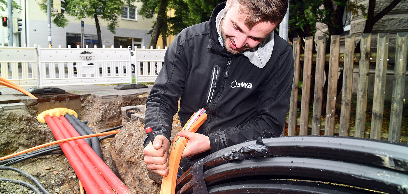 Arbeiter führt Kabelverlegung in einer offenen Baugrube durch: Im Vordergrund sind mehrere orangefarbene und schwarze Kabel sichtbar, die in die Erde eingebracht werden. Die Person trägt eine schwarze Jacke mit dem Logo ‚swa‘ und hält ein orangefarbenes Kabel in der Hand. Im Hintergrund sind Absperrungen und ein Holzzaun zu sehen, dahinter Bäume und Gebäude. Die Szene zeigt Arbeiten an der Energie- oder Telekommunikationsinfrastruktur.