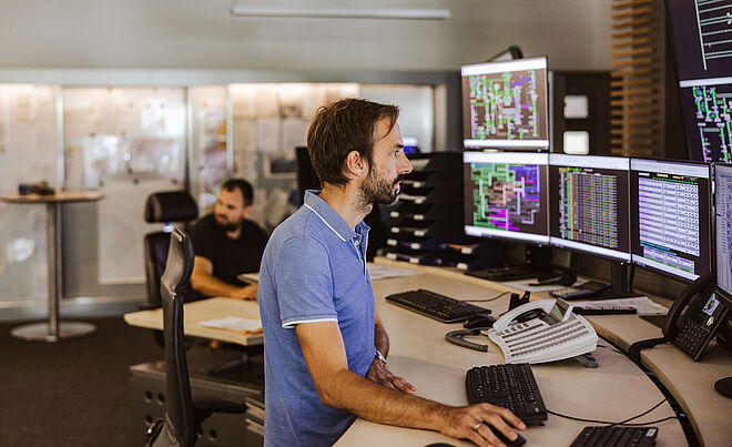 Mitarbeitende in einer modernen Leitstelle der swa Netze GmbH überwachen Energienetz‑Daten auf mehreren großen Monitoren. Eine Person sitzt an einem Arbeitsplatz mit Telefon, Tastatur und mehreren Bildschirmen, auf denen technische Netzvisualisierungen und Systemübersichten zu sehen sind. Im Hintergrund befindet sich ein weiterer Arbeitsplatz in einem hellen, offenen Kontrollraum. Verlinkung zur Auswahl Netzanschluss Seite.