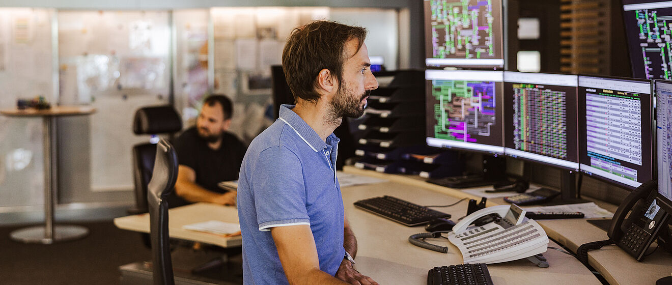 Mitarbeitende in einer modernen Leitstelle der swa Netze GmbH überwachen Energienetz‑Daten auf mehreren großen Monitoren. Eine Person sitzt an einem Arbeitsplatz mit Telefon, Tastatur und mehreren Bildschirmen, auf denen technische Netzvisualisierungen und Systemübersichten zu sehen sind. Im Hintergrund befindet sich ein weiterer Arbeitsplatz in einem hellen, offenen Kontrollraum. Verlinkung zur Auswahl Netzanschluss Seite.