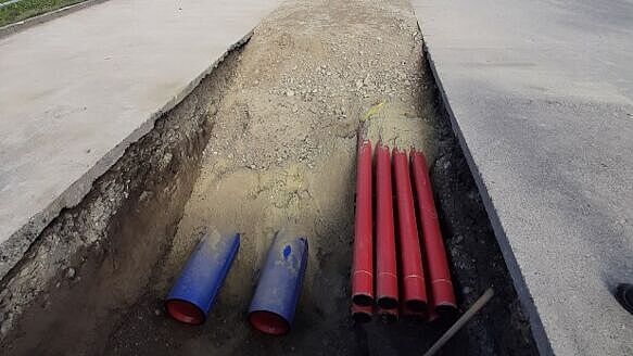 Baustelle für Leitungsverlegung auf einer Straße. Im Vordergrund ist ein geöffneter Graben mit mehreren roten und blauen Schutzrohren für Kabel oder Leitungen sichtbar. Der Graben verläuft entlang der Straße, die teilweise asphaltiert ist. Im Hintergrund stehen Baumaschinen, darunter ein Bagger und eine Rüttelplatte, sowie Absperrungen zur Sicherung der Baustelle. Links sind einige Bäume und rechts Gebäude zu erkennen. Die Szene zeigt Arbeiten zur Verlegung von Versorgungsleitungen.