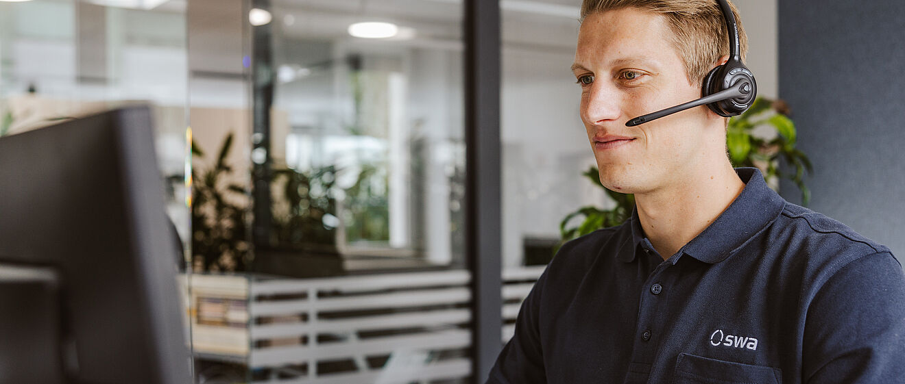 Eine Person sitzt an einem Schreibtisch und arbeitet an einem Computerbildschirm. Die Person trägt ein dunkles Poloshirt mit dem Logo „swa“ und ein Headset. Im Hintergrund sind Glaswände, Büroeinrichtung und einige grüne Pflanzen zu sehen. Die Szene zeigt eine moderne Büroumgebung, vermutlich in einem Kundenservice- oder Supportbereich.