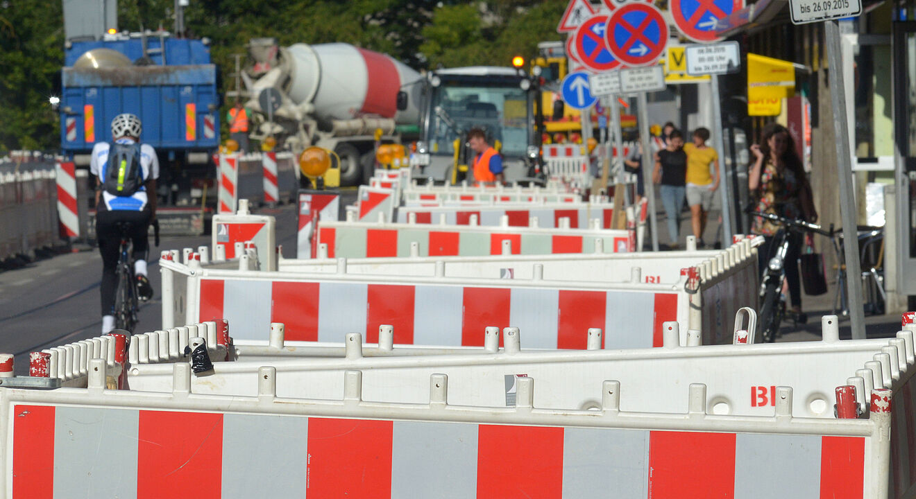 Eine Straßenbaustelle mit zahlreichen rot-weißen Absperrungen, die den Gehweg und die Fahrbahn trennen. Mehrere Verkehrsschilder sind sichtbar, darunter „Tempo 30“, „Fußgänger“, sowie mehrere Halteverbotsschilder. Im Hintergrund stehen Baustellenfahrzeuge, darunter ein Betonmischer und ein LKW. Auf der linken Seite fährt eine Person mit Fahrrad durch die Baustelle, während rechts Personen auf dem Gehweg gehen. Die Szene zeigt eine innerstädtische Baustelle mit umfangreichen Absperrungen und Verkehrshinweisen.