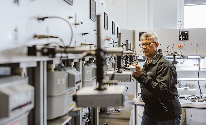 Prüfanlage für Gaszähler in einem technischen Raum. Mehrere Gaszähler sind in einer Reihe montiert und über Leitungen sowie Messgeräte verbunden. Eine Person in Arbeitskleidung mit ‚swa‘-Logo auf der Rückseite bedient die Anlage und überprüft die angeschlossenen Geräte. Die Prüfanlage besteht aus grauen Metallplatten mit Anzeigen, Anschlüssen und Halterungen für die Gaszähler. Verlinkung zur Zählerprüfstelle Seite.