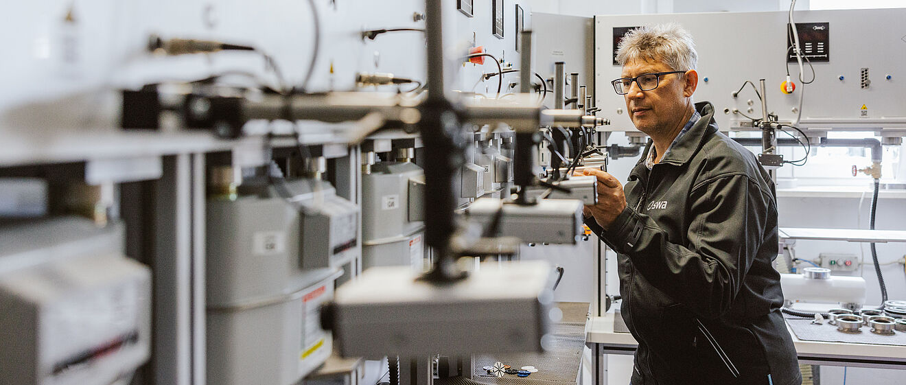 Prüfanlage für Gaszähler in einem technischen Raum. Mehrere Gaszähler sind in einer Reihe montiert und über Leitungen sowie Messgeräte verbunden. Eine Person in Arbeitskleidung mit ‚swa‘-Logo auf der Rückseite bedient die Anlage und überprüft die angeschlossenen Geräte. Die Prüfanlage besteht aus grauen Metallplatten mit Anzeigen, Anschlüssen und Halterungen für die Gaszähler. Verlinkung zur Zählerprüfstelle Seite.